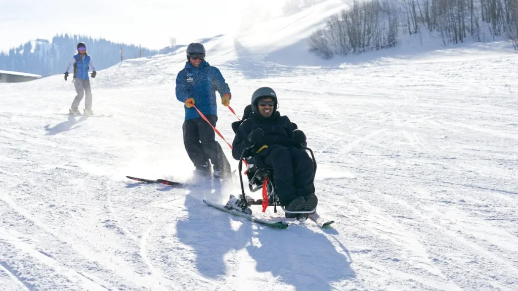An adaptive ski instructor guiding a skier in a sit-ski down a snowy mountain slope at the national ability centre, a popular activity for accessible winter travel.