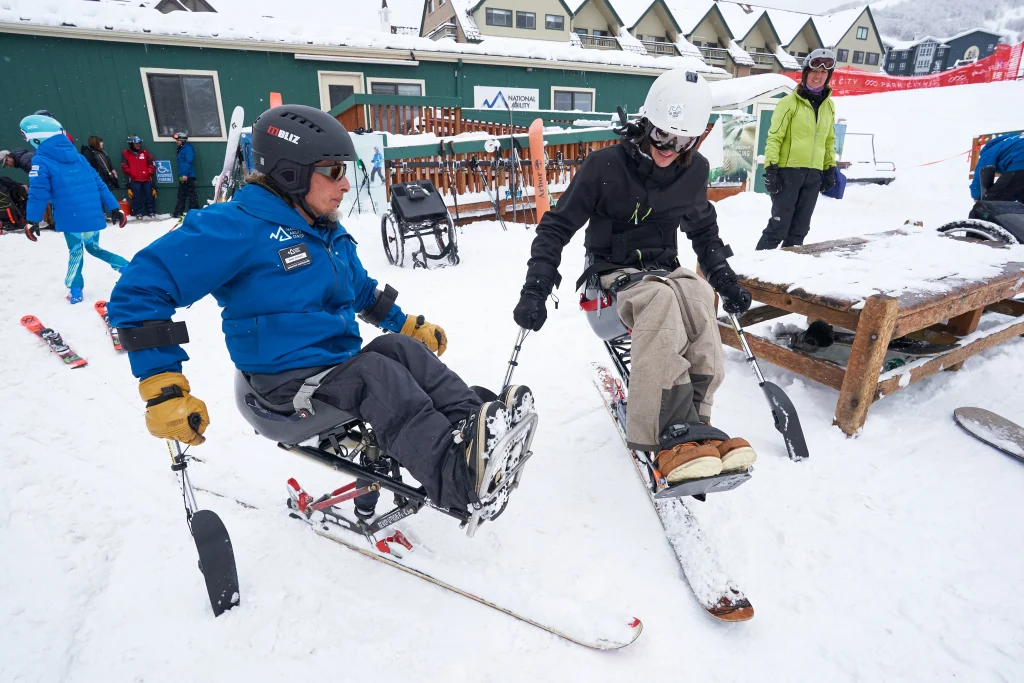 Two skiers using adaptive sit-skis preparing for a lesson on the snow, highlighting specialized equipment used in accessible winter travel.
